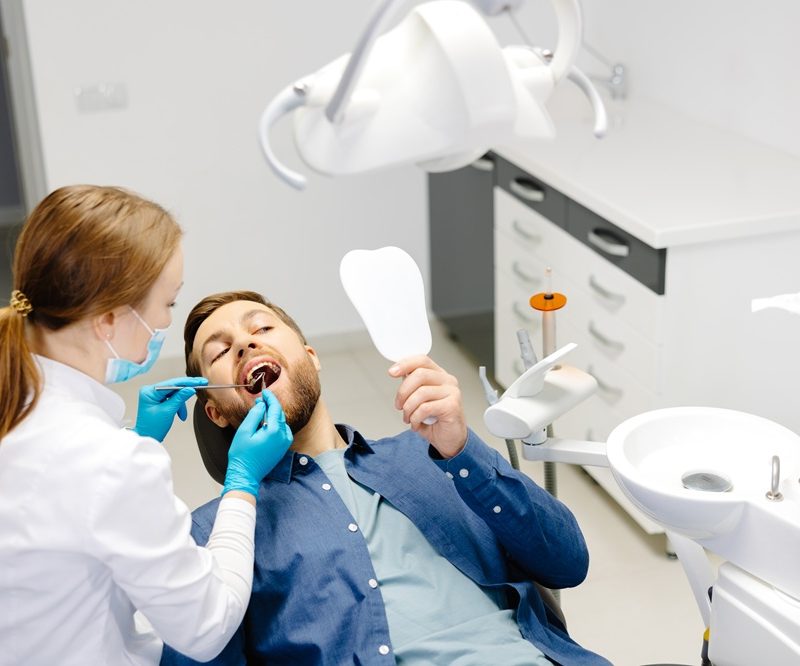 Dentist examining a patient’s teeth while the patient holds a mirror, helping detect early tooth decay symptoms during a routine dental visit – tooth decay symptoms